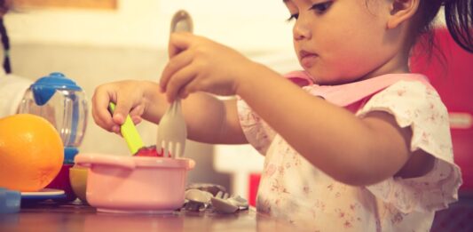 montre deux enfants jouant à la maîtresse et à l'élève dans une classe imaginaire. L'un des enfants tient un livre et pointe du doigt les lettres, tandis que l'autre enfant écoute attentivement. L'image illustre comment le jeu symbolique peut favoriser le développement du langage et des compétences de lecture.