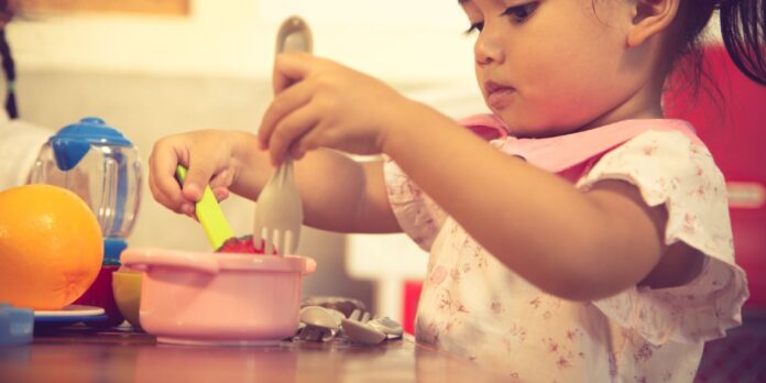 montre deux enfants jouant à la maîtresse et à l'élève dans une classe imaginaire. L'un des enfants tient un livre et pointe du doigt les lettres, tandis que l'autre enfant écoute attentivement. L'image illustre comment le jeu symbolique peut favoriser le développement du langage et des compétences de lecture.